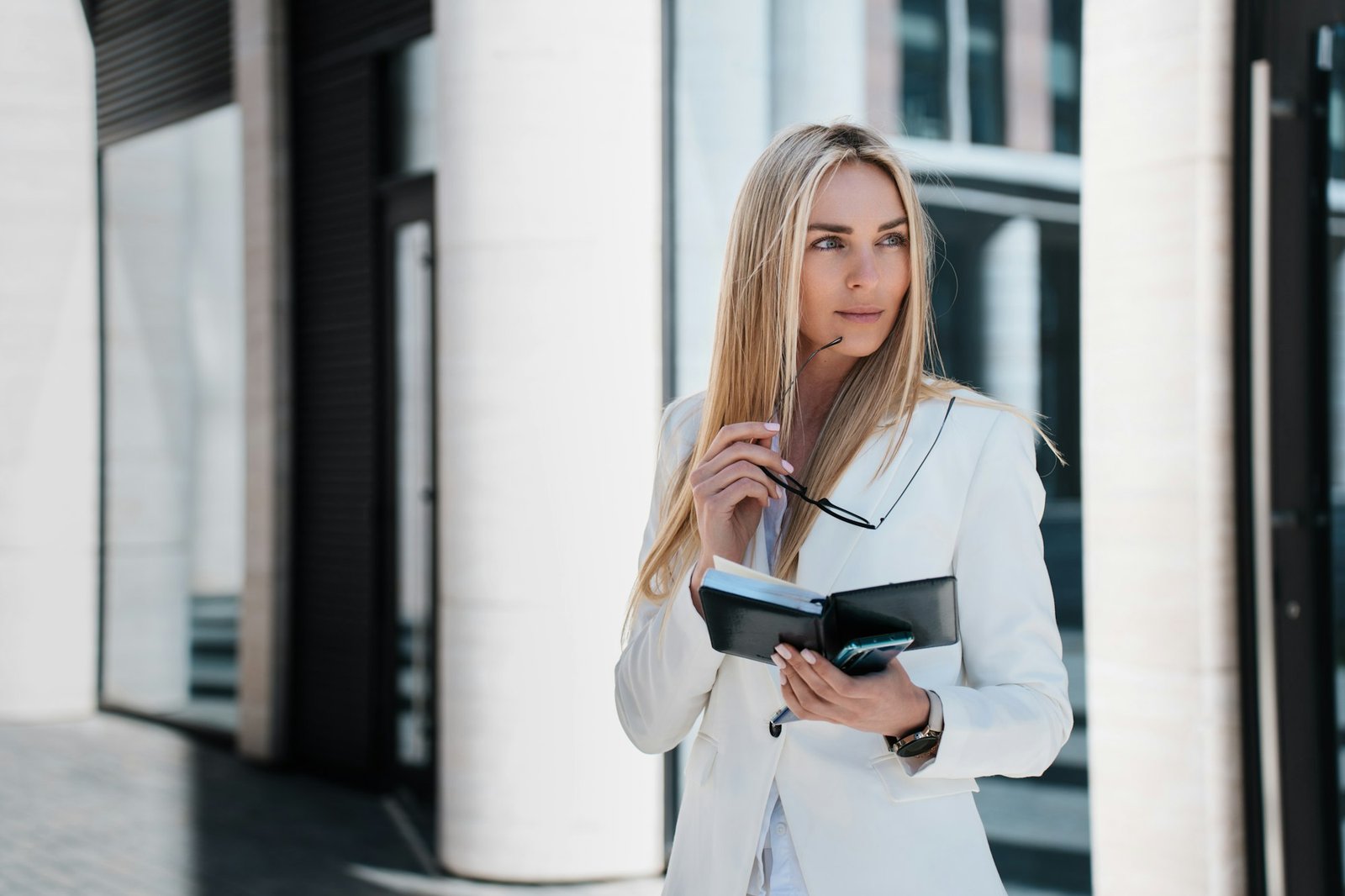 Attractive Swedish young woman lawyer in white suit outside, holds diary, spectacles, looks aside.